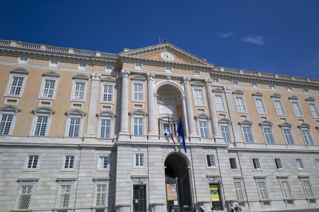 Main facade of the Royal Palace of Caserta with columns, flags and clear sky.